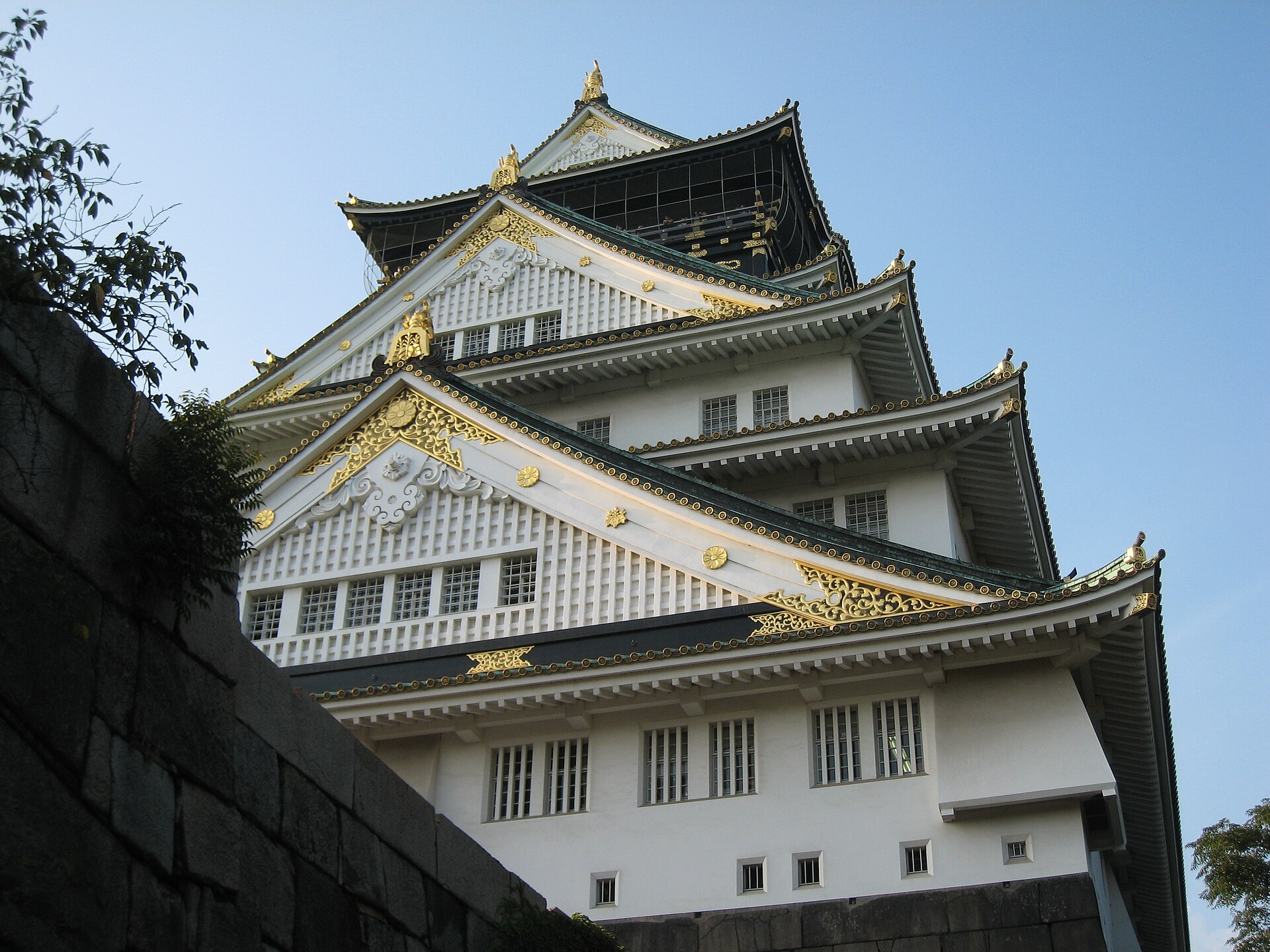 Donjon blanc et or du château d'Osaka, vu en contre-plongée sous un ciel bleu, entouré d'arbres.